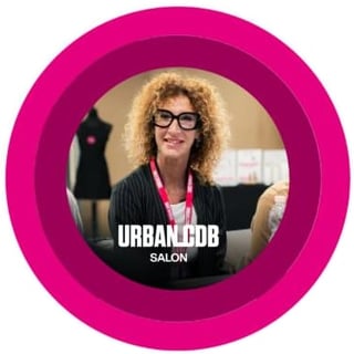 Woman with curly blonde hair and glasses wearing black jacket with pink lanyard in salon setting, UrbanCob Salon logo