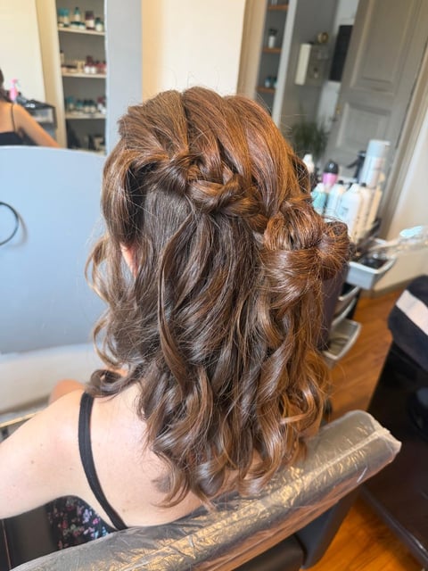 Back view of person with long brown wavy hair styled with a twisted crown braid, taken in a salon setting