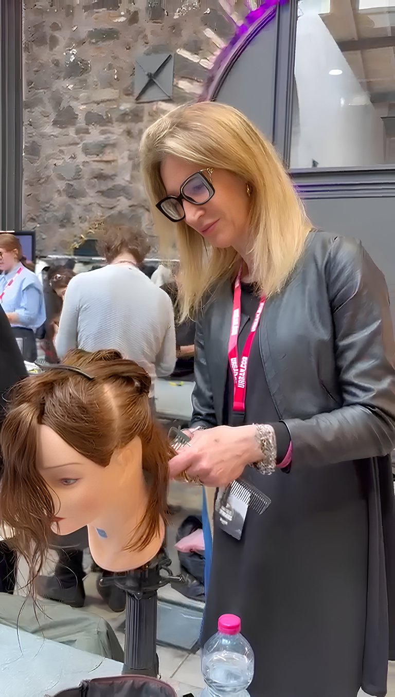 Hairstylist wearing glasses and ID badge styling hair on a mannequin head at an indoor salon or event venue