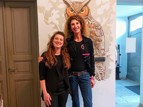 Two women posing together in front of a large owl artwork on a white wall, both wearing black clothing.